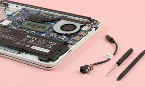 Professional studio photograph showing the internal components of an HP laptop with a replacement DC charging port cable, screwdriver, and prying tool on a light pink background for Atlantis repair services