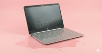 Professional studio photograph of a silver HP laptop sitting in a puddle of water with droplets on the screen and keyboard, set against a light pink background for Atlantis liquid damage repair services