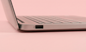 Professional studio photograph close-up of the side profile of a silver laptop, showing a USB port, set against a solid light pink background for Atlantis USB port repair services