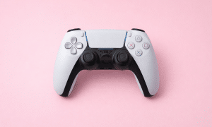 Professional 1:1 square studio photograph of a black PlayStation 4 DualShock controller and a white PlayStation 5 DualSense controller, set against a solid light pink background for Atlantis controller repair services.