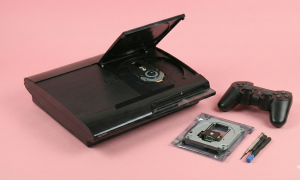 Professional studio photograph of a black Sony PlayStation 3 Super Slim console with its disc cover open, showing a replacement laser lens deck in a bag and repair tools beside it on a light pink background, representing Atlantis PS3 disc drive repair services.