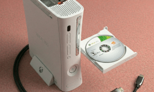 Close-up of an internal Xbox 360 optical disc drive and tray mechanism, positioned on a solid light pink background for Atlantis repair services.