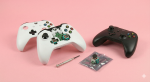 Professional studio photograph of white and black Xbox controllers on a light pink background; one white controller is partially disassembled showing internal analog stick modules, with a replacement joystick part and repair tools nearby for Atlantis repair services.
