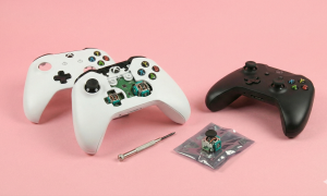 Professional studio photograph of white and black Xbox controllers on a light pink background; one white controller is partially disassembled showing internal analog stick modules, with a replacement joystick part and repair tools nearby for Atlantis repair services.