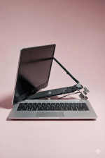Professional studio photograph of a silver HP laptop with a severely broken right hinge, showing the screen separated from the base with exposed metal components and wires, set against a light pink background.
