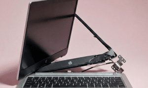 Professional studio photograph of a silver HP laptop with a severely broken right hinge, showing the screen separated from the base with exposed metal components and wires, set against a light pink background.