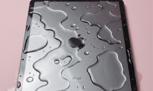 A professional technician using a digital microscope and precision micro-soldering tools to remove corrosion from an iPad Pro 2018 logic board, shown on a solid light pink background for Atlantis repair services.