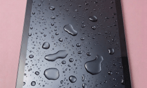 A professional technician using a digital microscope and precision tools to treat corrosion on an iPad logic board after a liquid spill, shown on a solid light pink background for Atlantis repair services.