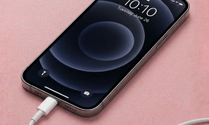 A professional technician replacing the internal Lightning connector and microphone flex cable assembly for an iPhone 12 Pro, displayed on a solid light pink background for Atlantis repair services.