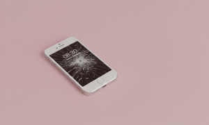 Professional 1:1 square studio photograph showing an Apple iPhone 4 with a shattered glass display, positioned on a solid light pink background for Atlantis repair services.