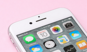 Professional 1:1 square studio photograph focusing on the top front assembly of an iPhone, showing the front-facing camera and ear speaker grille against a solid light pink background for Atlantis repair services.