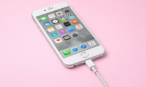 Professional 1:1 square studio photograph of a silver iPhone 7 with a white charging cable connected, set against a solid light pink background for Atlantis repair services.