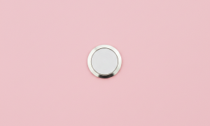 Professional 1:1 square studio photograph showing a close-up of the circular Home Button (Touch ID sensor) on the bottom bezel of a silver iPhone 7, set against a solid light pink background for Atlantis repair services.