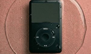 Close-up of a disassembled Apple iPod Classic logic board showing precision cleaning of the 30-pin dock connector and internal circuitry, positioned on a solid light pink background for Atlantis repair services.