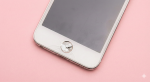 Close-up photograph of a white Apple iPod Touch displaying a severely cracked and shattered circular home button, set against a solid light pink background for Atlantis repair services