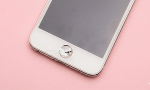 Close-up photograph of a white Apple iPod Touch displaying a severely cracked and shattered circular home button, set against a solid light pink background for Atlantis repair services