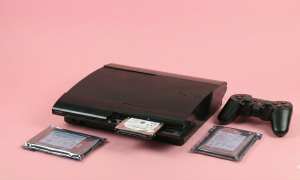 Professional studio photograph of a black PlayStation 3 Super Slim console with a 2.5-inch hard drive partially inserted into the drive bay, featuring replacement drives in anti-static packaging on a light pink background.