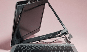 Professional studio photograph of a silver Sony laptop with a severely broken hinge and detached screen, showing exposed internal wiring and casing damage against a light pink background.
