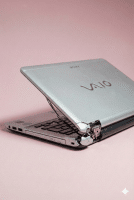 Professional studio photograph of a silver Sony Vaio laptop with a severely cracked rear casing and broken hinge, showing internal components through the damaged plastic, set against a light pink background.