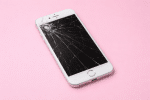 Professional 1:1 square studio photograph of a black iPhone 7 with a shattered and cracked glass screen, positioned on a solid light pink background for Atlantis repair services.