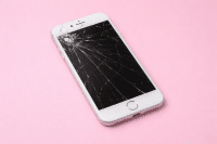 Professional 1:1 square studio photograph of a black iPhone 7 with a shattered and cracked glass screen, positioned on a solid light pink background for Atlantis repair services.