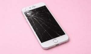 Professional 1:1 square studio photograph of a black iPhone 7 with a shattered and cracked glass screen, positioned on a solid light pink background for Atlantis repair services.