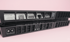 Professional 1:1 square studio photograph of the rear profile of a black PlayStation 4 console, highlighting the metal HDMI port against a solid light pink background for Atlantis console repair services.